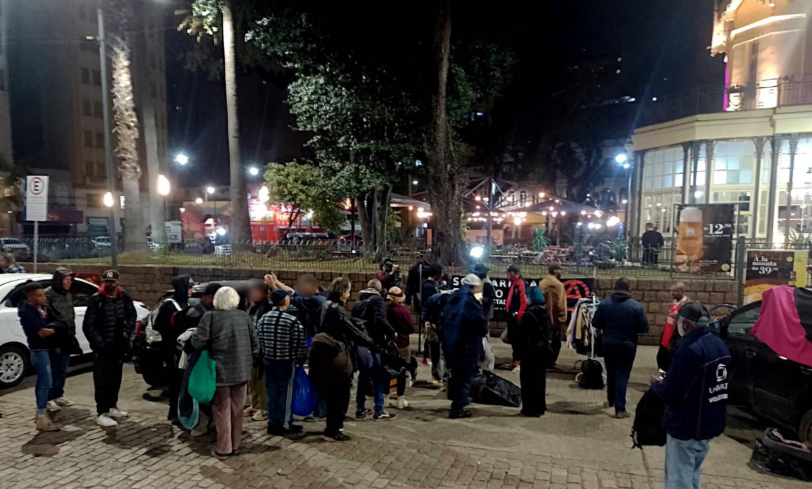 Foto de uma fila de pessoas esperando para acessar a distribuição de roupas. Ao fundo, o Chalé da Praça XV.