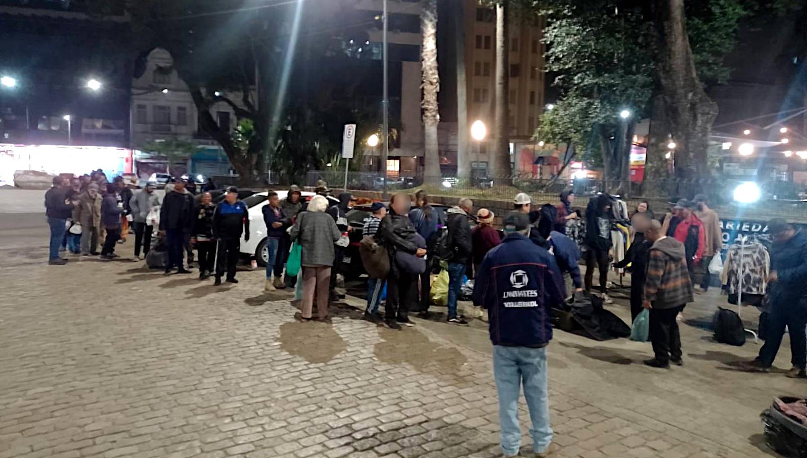 Foto mostra uma fila de dezenas de pessoas em uma praça aguardando para acessar a distribuição de roupas.
