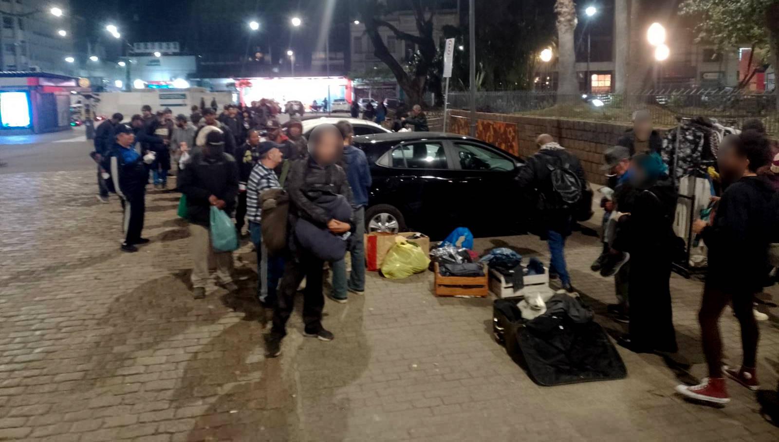 Foto mostra uma fila de dezenas de pessoas em uma praça aguardando para acessar a distribuição de roupas.