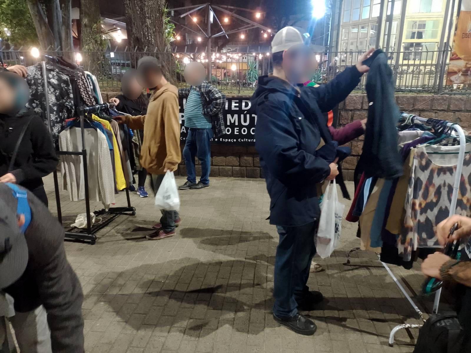 Foto de pessoas olhando e experimentando roupas em uma praça.