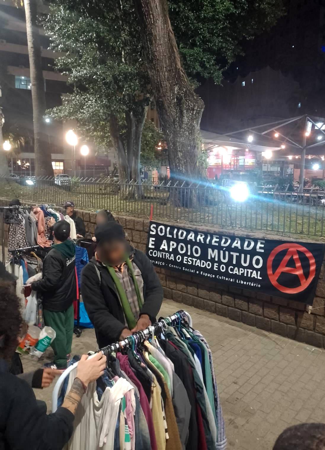 Foto. Em primeiro plano pessoas olhando roupas penduradas em três araras. Ao fundo uma faixa de tecido preto pendurada em um muro de pedra. Na faixa está escrito em letras garrafais brancas: "Solidariedade e apoio mútuo contra o Estado e o Capital." Abaixo em letras menores, está escrito: "Espaço - Centro Social e Espaço Cultural Libertário". E ao lado um grande A vermelho dentro de um círculo.