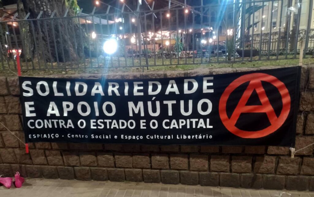 Foto de uma faixa de tecido preto pendurada em um muro de pedra. Na faixa está escrito em letras garrafais brancas: "Solidariedade e apoio mútuo contra o Estado e o Capital." Abaixo em letras menores, está escrito: "Espaço - Centro Social e Espaço Cultural Libertário". E ao lado um grande A vermelho dentro de um círculo.