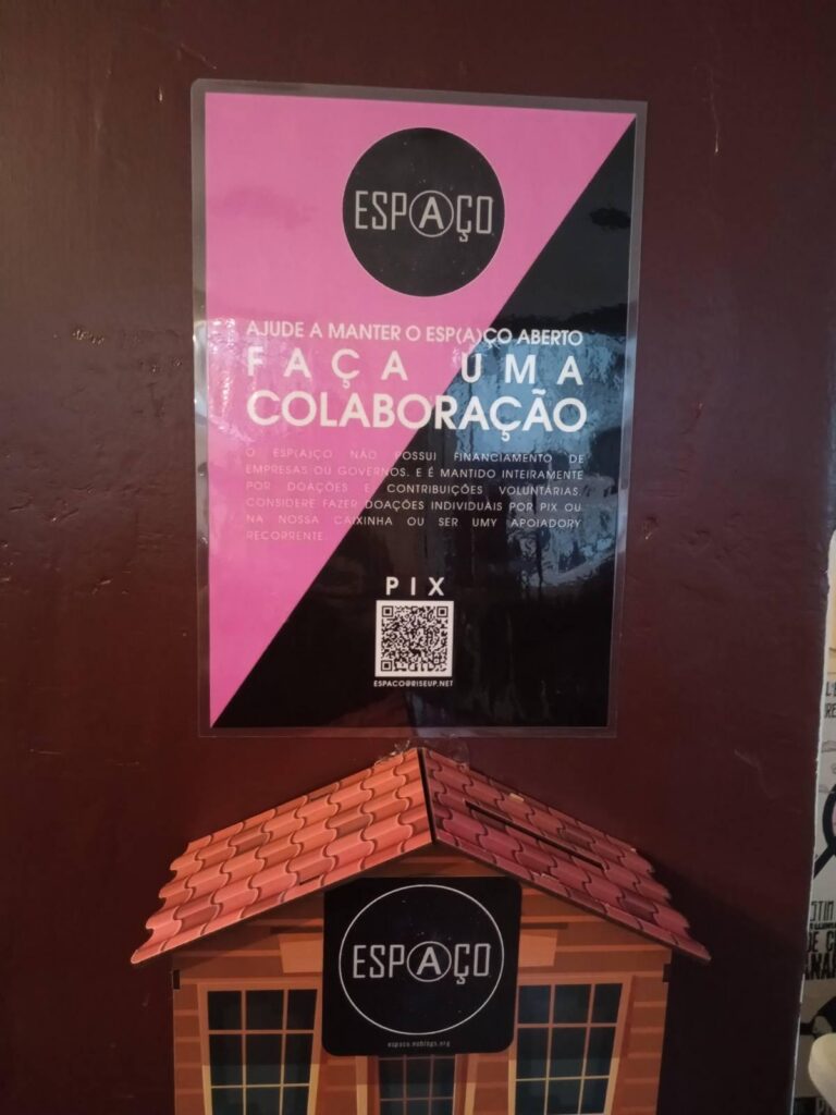 Foto de um cartaz colado na parede: dividido na diagonal nas cores rosa e preto. No topo o logotipo do espaço e abaixo escrito: "Ajude e manter o Esp(a)ço aberto: Faça uma colaboração". Abaixo do cartaz um cofrinho em formato de casa com um adesivo do Espaço colado na fachada.
